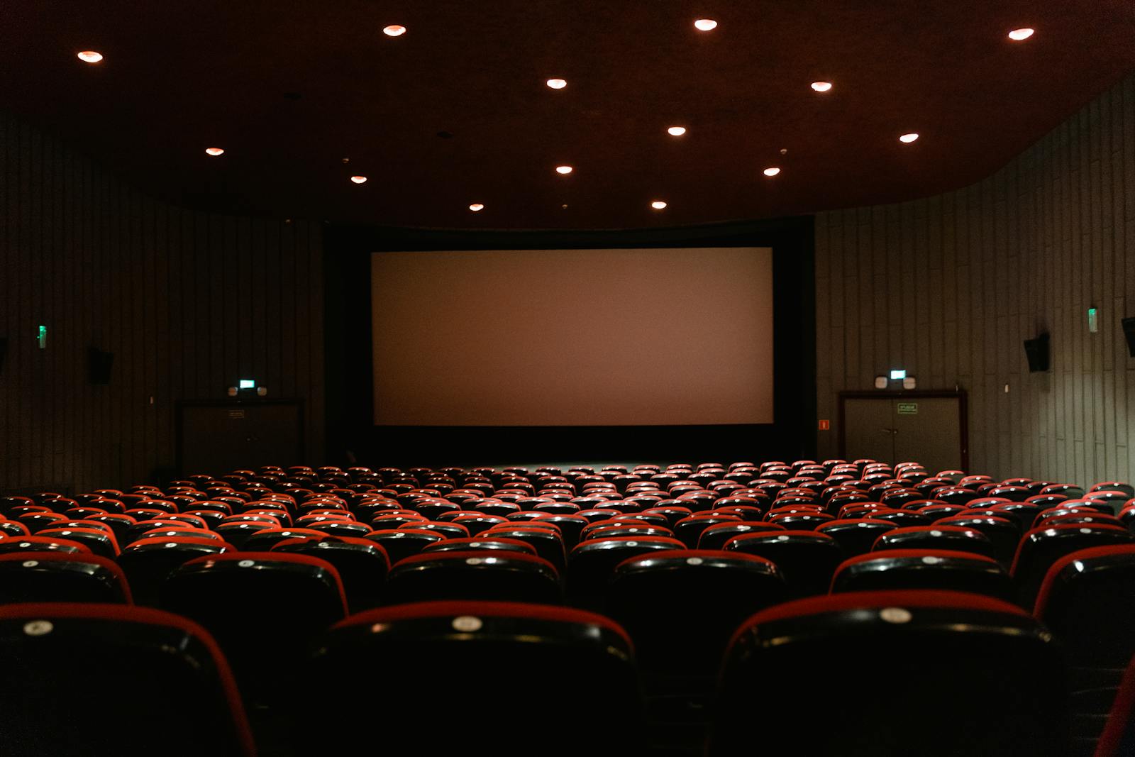 A dimly lit empty cinema theater with rows of red chairs facing a projector screen.